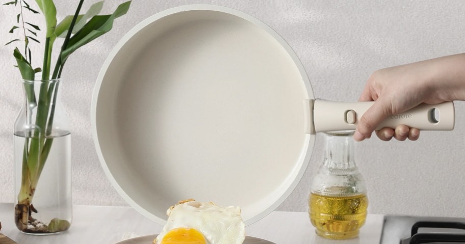 A hand holding a cream-colored Carote nonstick frying pan with a detachable handle, tilting it to slide a fried egg onto a plate.