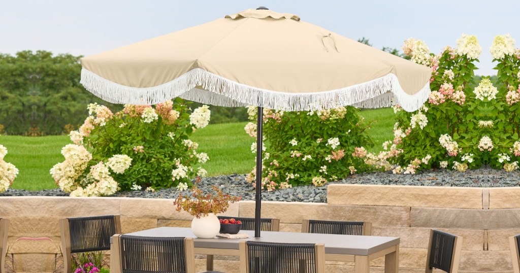 Cream colored umbrella with white fringe set up with patio table outside on stone patio