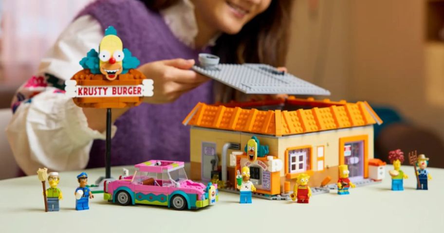 A woman playing with a Simpsons Krusty Burger LEGO set on a coffee table