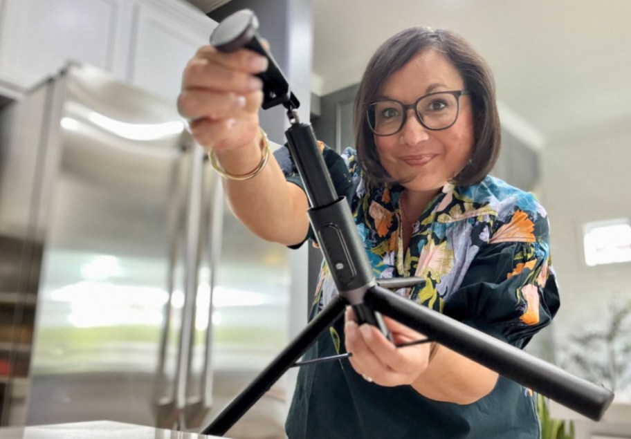 woman holding her phone tripod, showing it to the camera while standing in the kitchen