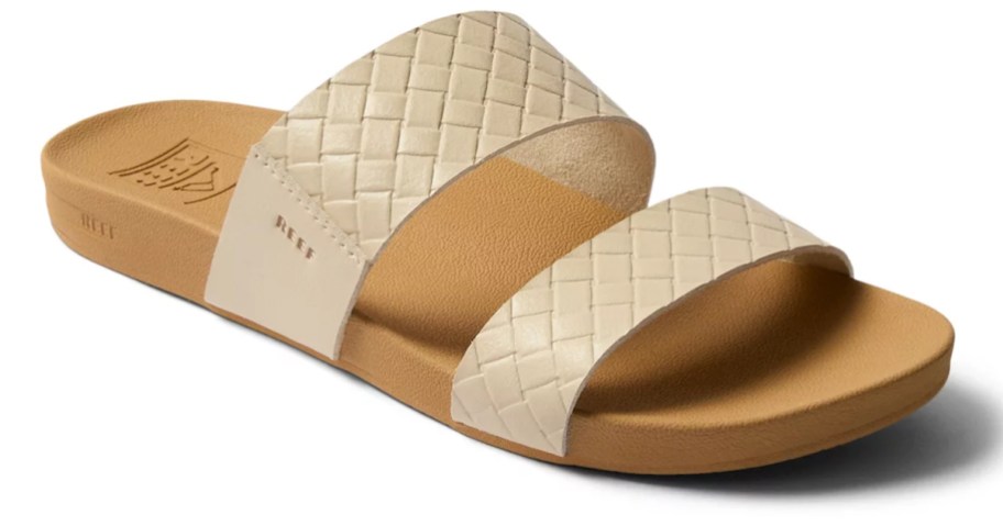 A REEF Kaia slide sandal featuring two wide woven cream-colored straps and a tan cushioned footbed with subtle branding on the side.