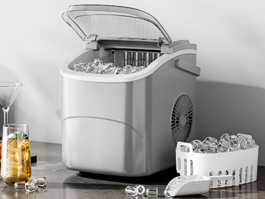 A silver ice maker on the counter surrounded by beverages and ice 