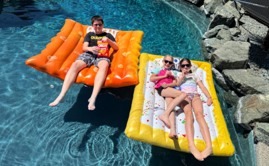 a boy laying on an orange cheezit pool float next to a poptarts pool float with two girls, all in a pool with a rock border