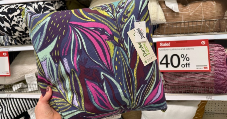 hand holding up a colorful leaf print outdoor pillow, sign on shelf behind it says "40% off"