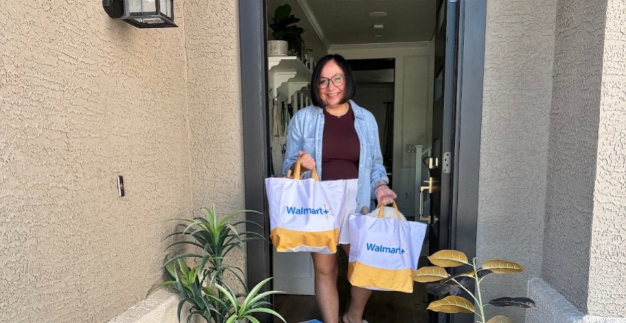 smiling woman standing her in open doorway holding two walmart plus delivery bags