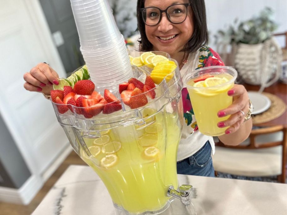 woman standing behind a Buddeez Drink Dispenser with fresh strawberries, lemons, limes, and plastic cups on top in the party lid and the inside filled with lemonade, while holding a plastic cup of lemonade