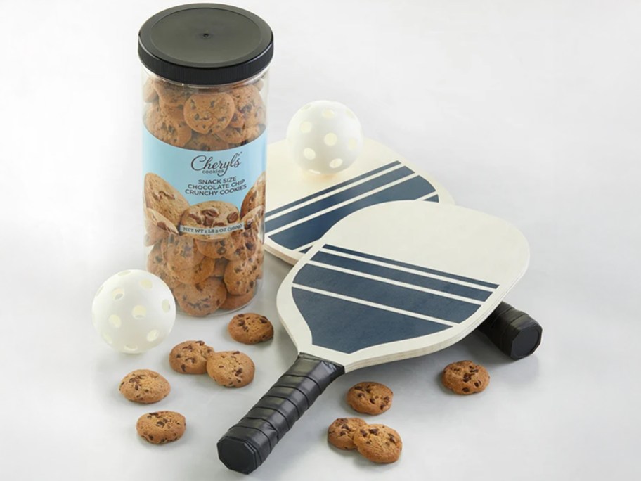 Cheryl's Pickleball gift set featuring two white paddles with navy stripes, two perforated white balls, and a tall container of snack-size chocolate chip cookies scattered around.