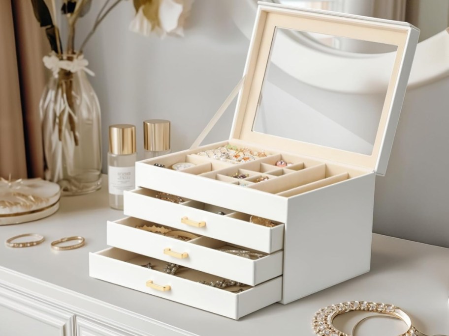 a large white jewelry box with 3 drawers and a glass top on a dresser, the drawers and top are open