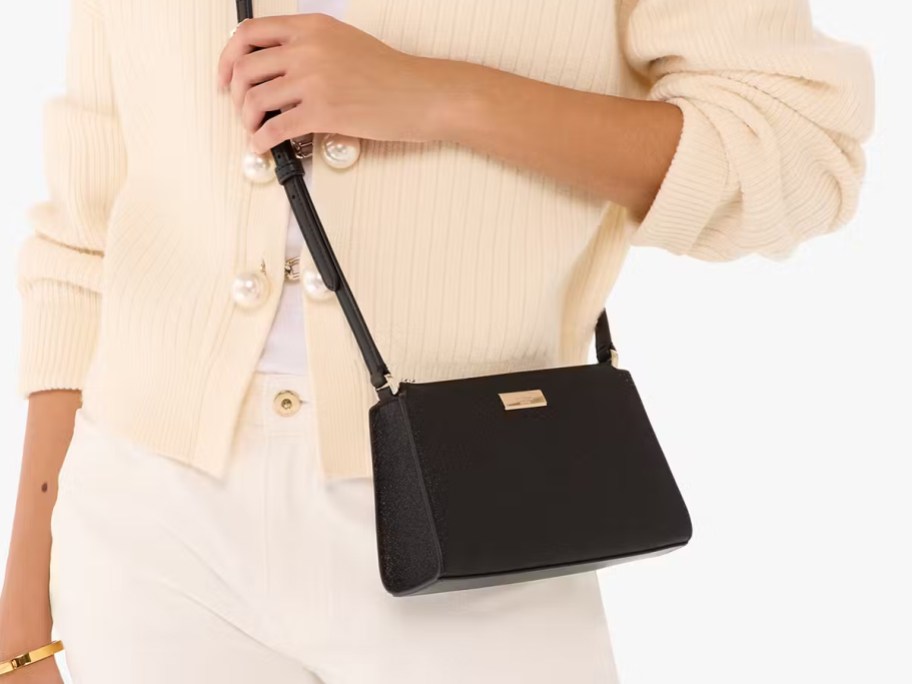 Woman wearing a cream cardigan with pearl buttons and white pants, carrying a black Limited Edition Glitter Mini Crossbody bag with a slim adjustable strap.