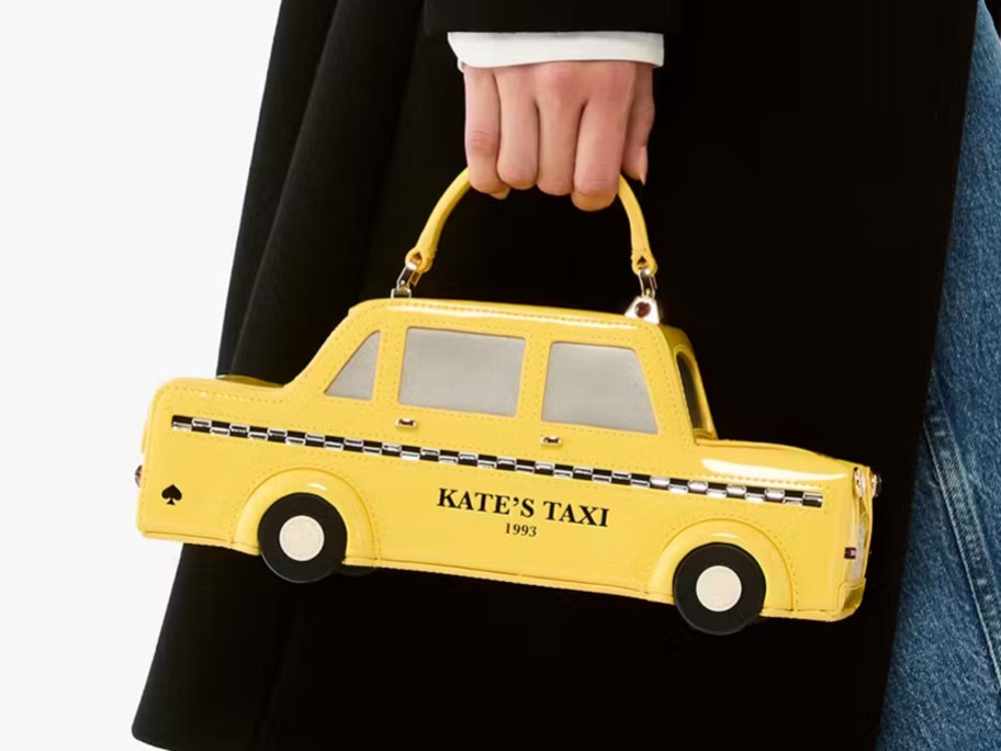 Person holding a Kate Spade New York 3D taxi crossbody bag shaped like a yellow cab with "KATE'S TAXI 1993" printed on the side.