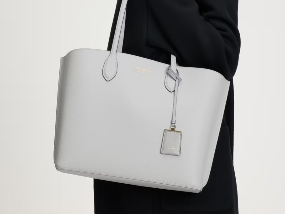 Person carrying a light gray Kate Spade Suite Large Work Tote with top handles and a matching luggage tag charm.