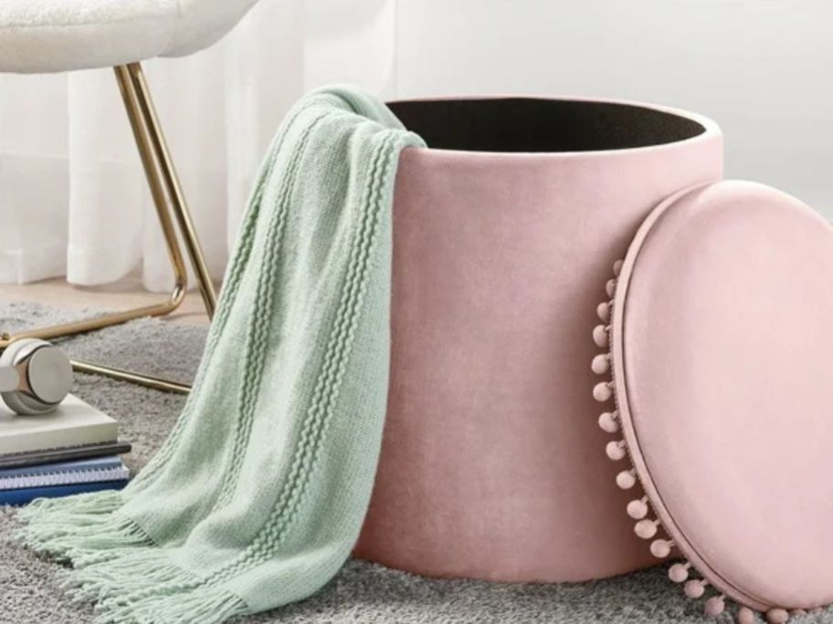 A pink ottoman with pom poms around the top holding a blanket