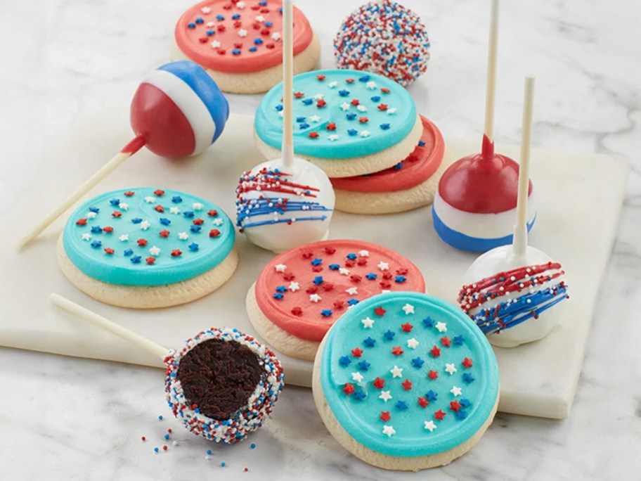 A festive assortment of patriotic treats featuring red, white, and blue cake pops decorated with sprinkles and drizzle, alongside sugar cookies topped with colorful frosting and star-shaped sprinkles.