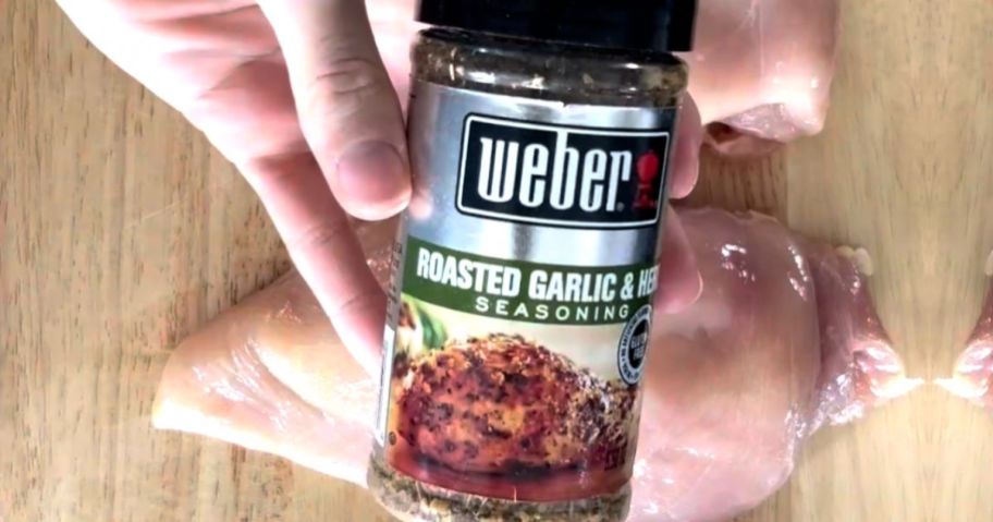 Hand holding a bottle of Weber Roasted Garlic & Herb Seasoning over raw chicken breasts