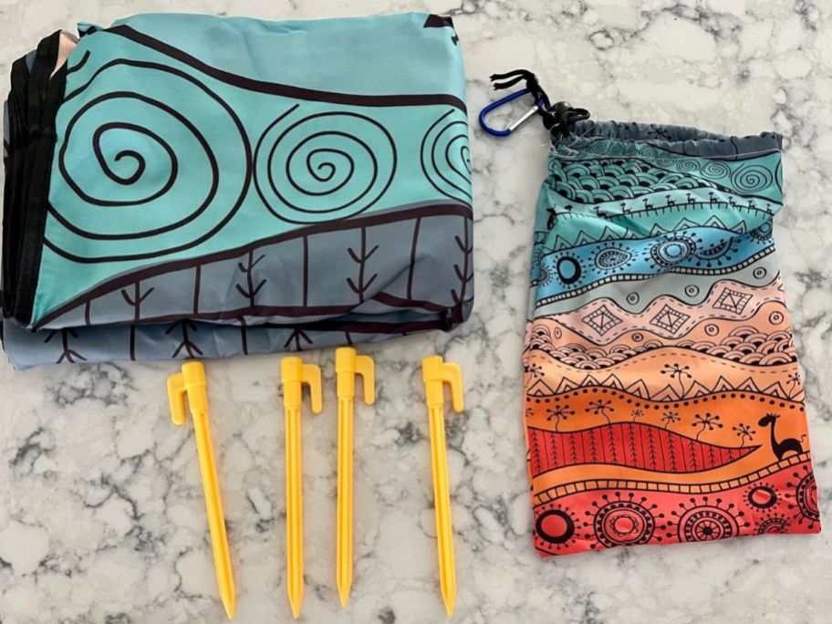 blue and orange Waterproof Beach Blanket folded up with four yellow stakes and bag with carabiner next to it on table