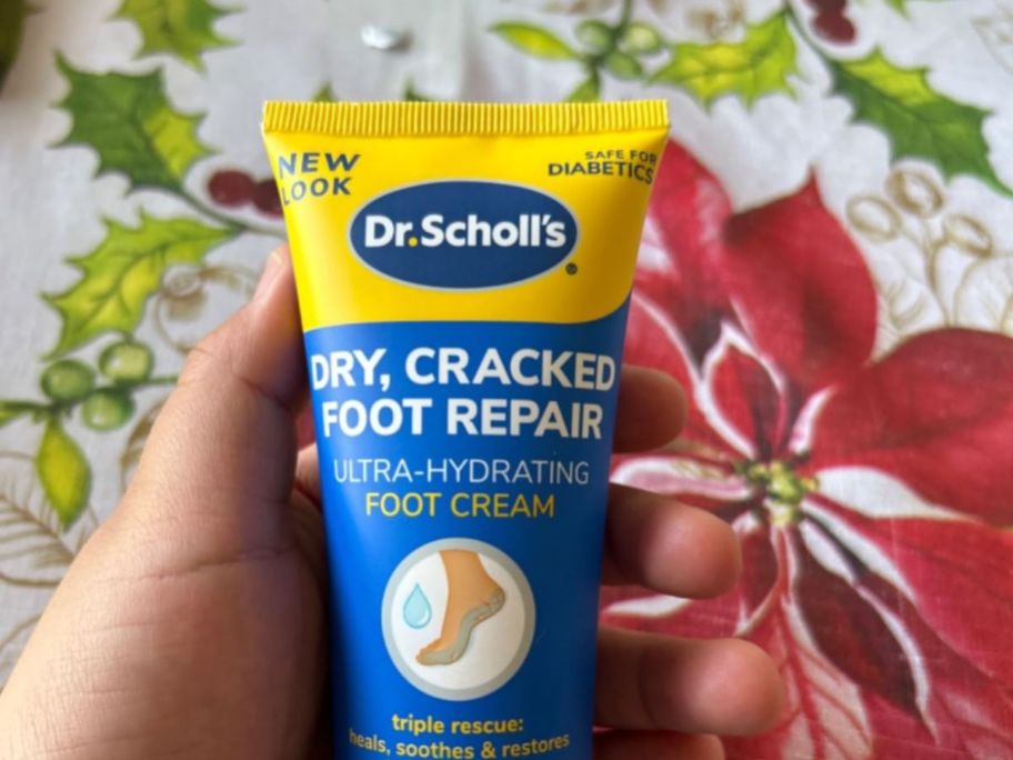 blue and yellow bottle of Dr. Scholl's Foot Cream in hand above floral tablecloth