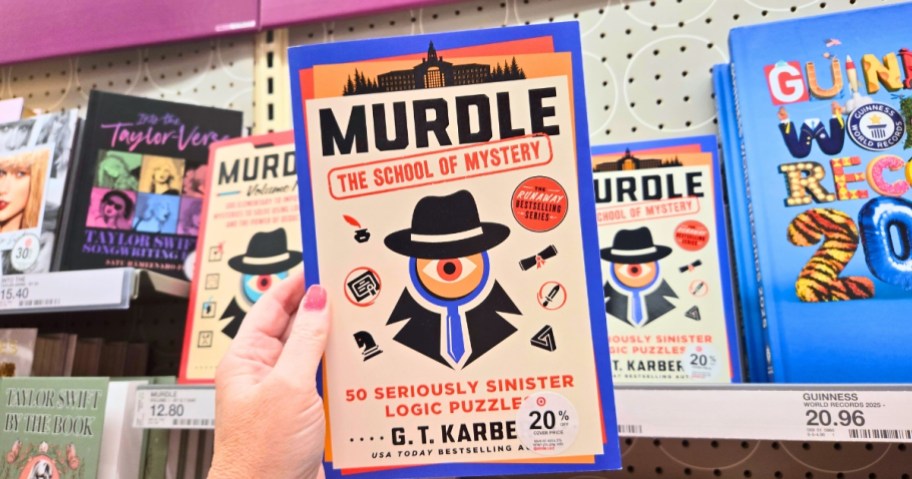 hand holding a Murdle mystery puzzle book with more on a store shelf behind it