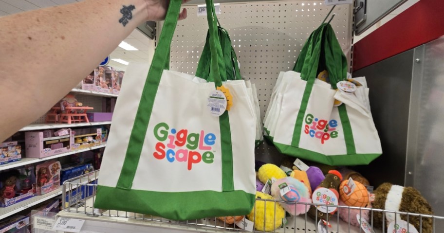 hand holding a white and green Gigglescape tote bag, more on a store rack behind it